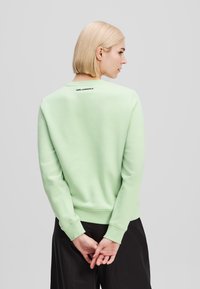 Felpa verde chiaro con polsini e orlo a coste. Presenta un collo rotondo e un logo stampato in nero nella parte posteriore. Texture morbida e vestibilità rilassata.