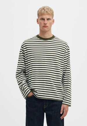 Un homme blond porte une chemise oversize à manches longues, rayée de blanc et de vert foncé, avec un jean foncé, sur un fond clair uni.