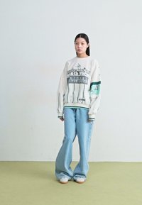 Λευκή υπερμέγεθη προπονητική μπλούζα με γραφικό μοτίβο και πράσινες λεπτομέρειες, συνδυασμένη με ανοιχτό μπλε παντελόνι φάρδους και λευκά αθλητικά παπούτσια.