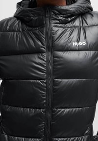 Černá péřová bunda s prošívaným vzorem, na zip a s kapucí. Na levé straně hrudi má výrazné bílé logo "HUGO".