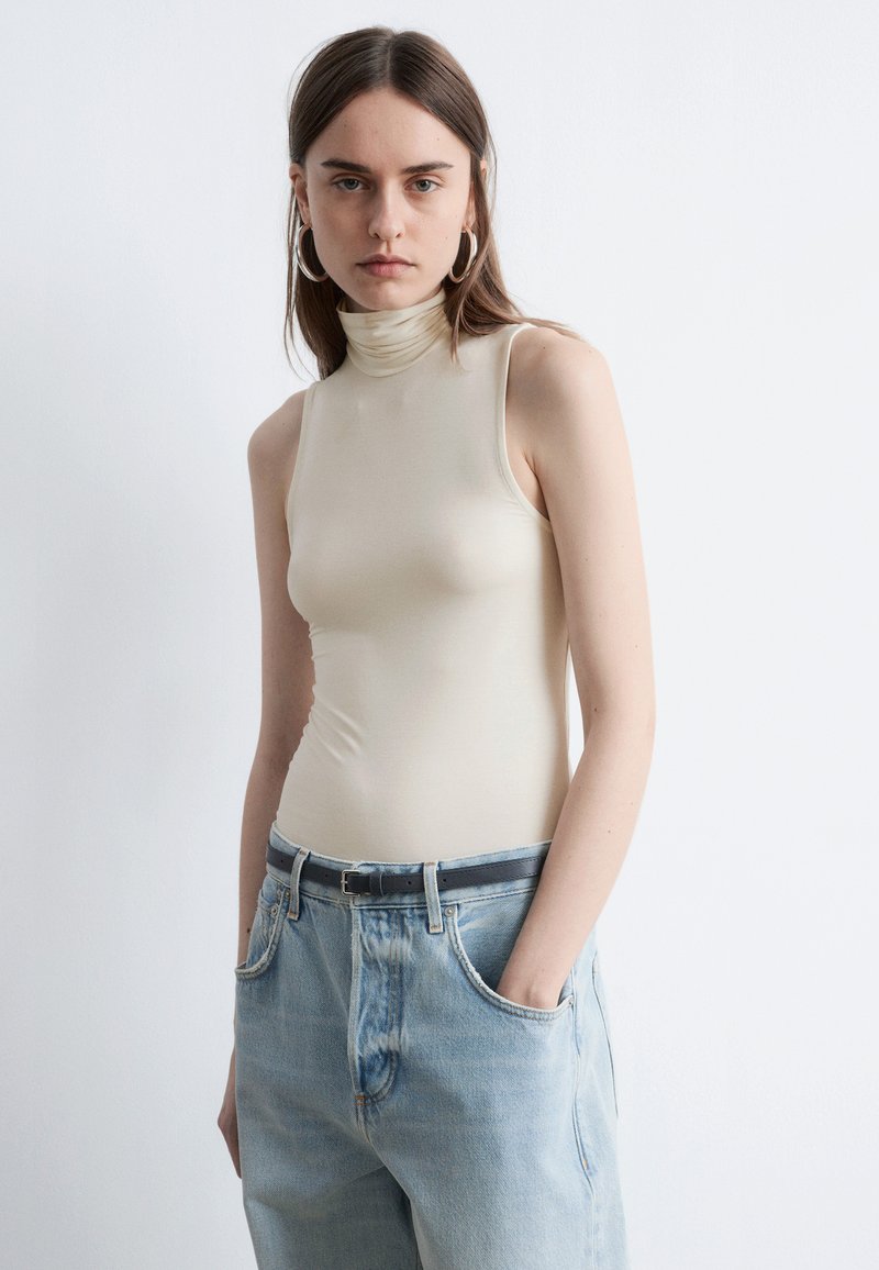 Haut beige sans manches à col roulé en tissu lisse, associé à un jean large délavé clair, avec une ceinture noire et de grandes boucles d'oreilles.