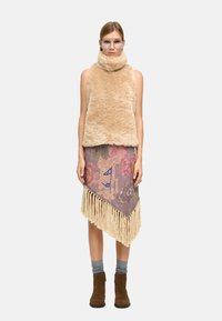 Beżowa kamizelka z sztucznego futra bez rękawów, zestawiona z frędzlami i wzorzystą spódnicą w pastelowych kolorach. Modelka ma na sobie szare skarpetki i brązowe botki za kostkę.