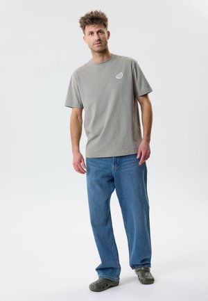 Mann steht, trägt ein graues T-Shirt mit Zitronenscheiben-Logo, lockere blaue Jeans und olivgrüne Crocs vor weißem Hintergrund.