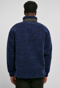 Dunkelblauer Fleece-Pullover mit strukturierter Stoffoberfläche, hohem Kragen und einem schwarzen Nylon-Element am Hals. Verfügt über eine lockere Passform und lange Ärmel.