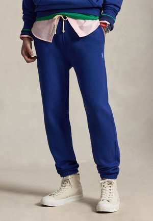 Partie inférieure portant un pantalon de survêtement bleu avec des poignets élastiques, des baskets montantes beiges et des hauts superposés bleus, verts et roses avec une montre rouge visible.