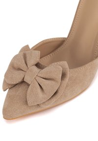 Zapato pump de ante beige con tacón alto, punta puntiaguda y un gran lazo simétrico decorativo en el empeine. Forro interior liso visible.