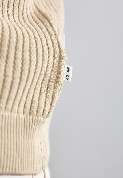 Suéter de punto acanalado color beige con cuello redondeado y una pequeña etiqueta en el dobladillo. La textura es suave y el diseño es sencillo.