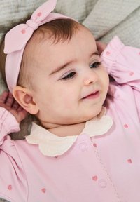 Helles rosa Overall mit kleinen Herzmustern, ausgestattet mit einem weißen Spitzenkragen und einem rosa Haarband mit Schleife. Weicher Stoff und lockere Passform.