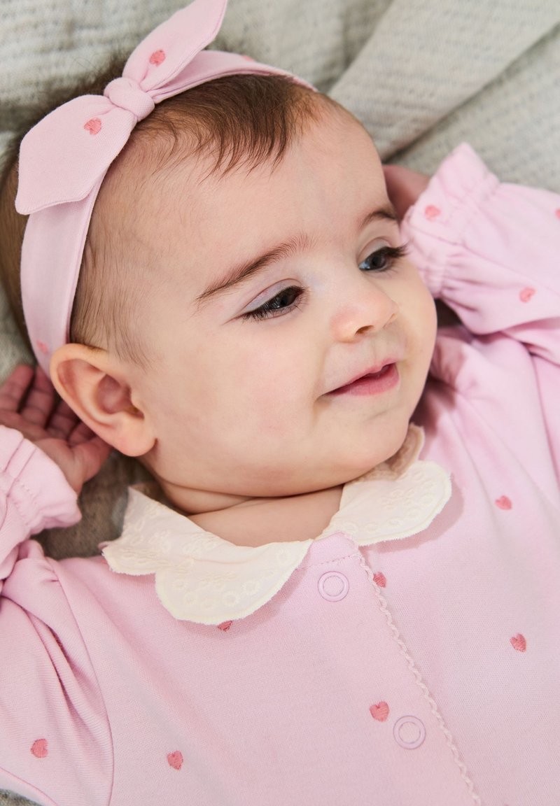 Helles rosa Overall mit kleinen Herzmustern, ausgestattet mit einem weißen Spitzenkragen und einem rosa Haarband mit Schleife. Weicher Stoff und lockere Passform.