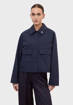 Femme portant une veste bleu marine boutonnée avec de grandes poches à rabat et un pantalon large sombre à motifs, debout devant un fond clair uni.