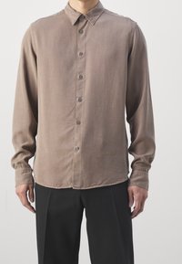 Camisa de manga larga en tela marrón claro, con cuello abotonado, botones en la parte frontal y puños fruncidos, con un detalle sutil del logo en el dobladillo.