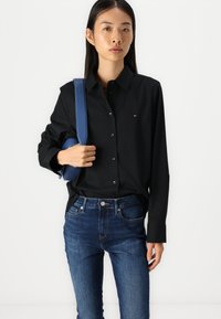 Zwarte blouse met lange mouwen, ontworpen om slim te passen. Marineblauwe schoudertas met een gladde textuur. Donkerblauwe spijkerbroek.
