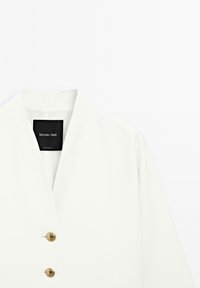 Veste blanche avec un design en V, ornée de boutons dorés, style minimaliste et texture lisse. Étiquette visible à l'intérieur du col.