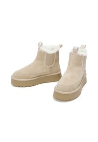 Stivali alla caviglia in suede beige con fodera in pelliccia sintetica, pannelli elastici laterali e una robusta suola in tan. Include linguette per una facile indossabilità.