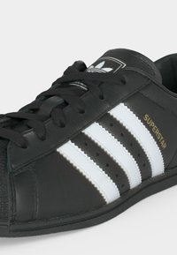 Schwarzer Adidas Superstar Sneaker mit weißen Streifen, schwarzen Schnürsenkeln und goldfarbenem "SUPERSTAR"-Schriftzug an der Seite.