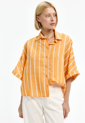 Frau trägt ein lockeres orangefarbenes Hemd mit weißen vertikalen Streifen und weiße Hose, schaut nach links vor weißem Hintergrund.