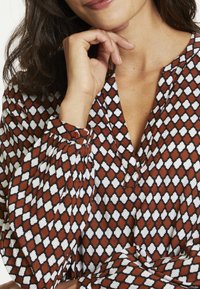 Blusa a maniche lunghe con un motivo geometrico rosso e bianco su uno sfondo scuro, con polsini abbottonati e scollatura a V.