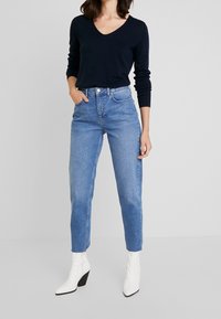 Marine V-hals trui, blauwe high-waisted jeans en witte puntige enkel laarzen met dikke hakken. Glad textuur, casual ontwerp.