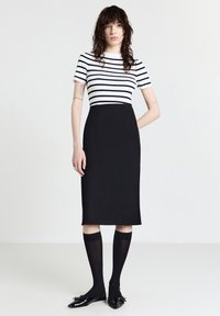 Frau mit welligem braunem Haar, die ein weiß-gestreiftes Oberteil, einen schwarzen knielangen Rock, schwarze kniehoche Socken und schwarze spitze Ballerinas mit Schleifen trägt.