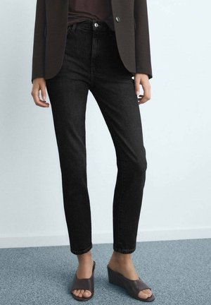 Person iført sorte skinny jeans med høj talje, brun blazer og grå sandaler med kilehæl og åbne tæer, stående på gråt tæppe.