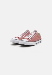 Zapatillas de lona rosas con cordones blancos, punteras de goma y rayas negras a lo largo de la suela. Presentan un diseño clásico de corte bajo.