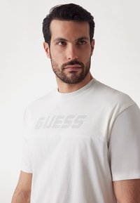 White T-shirt featuring a mesh design at the top section, with a smooth texture and printed logo. Short sleeves and a crew neckline.