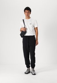 Witte katoenen T-shirt met een logo op de borst, gecombineerd met zwarte sweatpants. Een kleine zwarte crossbody tas en grijze sneakers maken de look compleet.