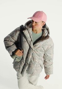 Vrouw draagt een gewatteerd jasje met patroon, een roze baseballpet, grijze broek en draagt een kleine groene tas over één schouder.