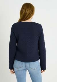 Sudadera acolchada azul marino con cuello redondo y mangas largas, con un patrón texturizado. Combinada con vaqueros ajustados de color azul claro.