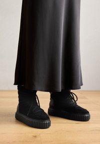 Svarte høye sneakers med teksturert stoff, flate lisser og tykk skråsåle, sammen med en lang, flytende svart skjørt.