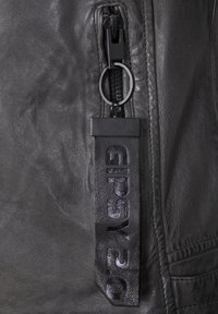 Detail einer schwarzen Lederjacke mit einem Reißverschluss und einem runden Ziehgriff sowie einem geprägten Stoffetikett mit "GIPSY 20" in dunkler Schrift.