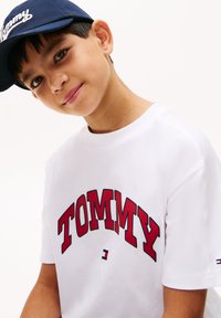 Hvit bomulls T-skjorte med fet rød "TOMMY" tekst som buer over brystet. Korte ermer med logo detaljer på ermene.