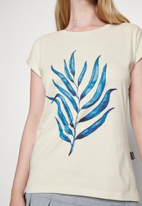 T-shirt de manga curta em creme com um design de folha azul, em tecido suave e com um corte descontraído. Inclui uma pequena etiqueta da marca na lateral.