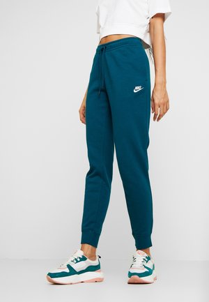 Vrouw draagt turquoise Nike joggingbroek met een witte cropped top en turquoise, witte en roze sneakers, staand tegen een effen lichte achtergrond.