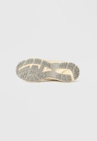 Gaiši brūna sporta apavi zolīte ar teksturētu pelēku gumijas protektoru, kurā redzams izteikts arkveida izliekums un dažādi saķeres raksti, nodrošinot labu saķeri.