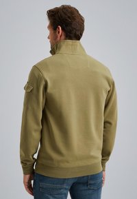 Olive zielony sweter z wysokim kołnierzem, wykonany z miękkiego materiału. Posiada kieszeń na ramieniu oraz ściągacze z gładką fakturą.