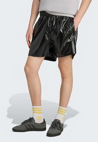 Svarta, glansiga träningsshorts med elastisk midja och sidofickor, kombinerade med grå sneakers och strumpor med gula ränder.