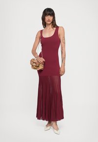 Abito senza maniche color bordeaux con top aderente e gonna plissettata, abbinato a una borsa intrecciata multicolore e scarpe bianche con punta.