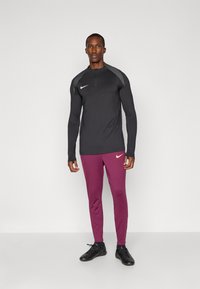 Top desportivo de manga longa preto com um design cinza nos ombros, combinado com umas calças ajustadas cor bordeaux. Ambos apresentam o logo da Nike. Sapatos pretos.