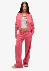 Conjunto de chándal rosa que incluye una chaqueta con cremallera y pantalones con cordón. Material de algodón suave con puños elásticos y un logo impreso en la parte superior.