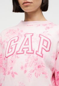 Γυναίκα που φοράει ανοιχτό ροζ φούτερ με ροζ λουλουδάτο σχέδιο και έντονο λογότυπο GAP στο στήθος, κομμένο στο ύψος του στόματος και του λαιμού.