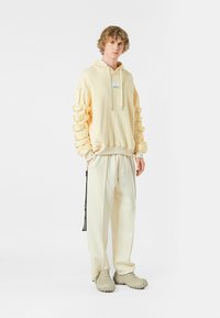 Sudadera amarilla con capucha y diseño de mangas en capas, combinada con pantalones crema y calzado texturizado beige. Presenta un parche de logo en el frontal.
