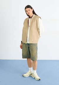 Chaleco sin mangas beige sobre un suéter blanco, combinado con pantalones cortos de mezclilla y zapatillas deportivas verde claro; diseño simple con detalles de hardware mínimos.