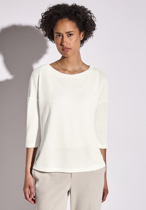 Witte, gestructureerde top met driekwartmouwen en een ronde halslijn, gedragen over lichtbeige broeken, met een relaxte pasvorm en subtiele glans.