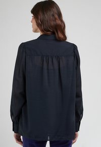 Blusa blu navy con collo a punta, maniche lunghe e voluminose, e dettagli posteriori plissettati. Tessuto liscio con una consistenza morbida e leggera.
