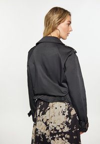 Cappotto trench corto nero realizzato in tessuto liscio, con colletto classico e polsini con cintura, abbinato a una gonna a fantasia floreale.