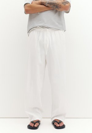 Person med tatoverede arme krydsede, iført hvide bukser med snøre, sorte sandaler og en stribet kortærmet skjorte, stående mod en ensfarvet baggrund.