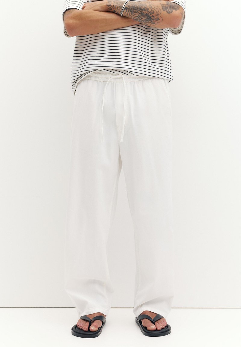 Personne aux bras tatoués croisés, portant un pantalon blanc à cordon, des sandales noires et une chemise rayée à manches courtes, debout devant un fond uni.