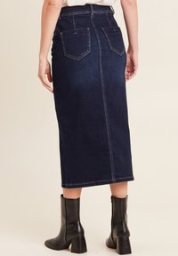 Gonna midi in denim scuro con vita aderente, tasche posteriori e cuciture a contrasto. Indossata con stivali neri con tacco. Design semplice e classico.