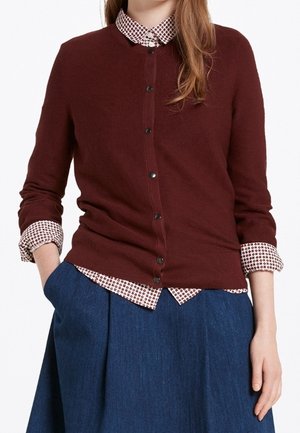 Maglione bordeaux con bottoni frontali, texture a costine e maniche lunghe, sovrapposto a una camicia bianca a quadri con colletto fantasia. Gonna blu.